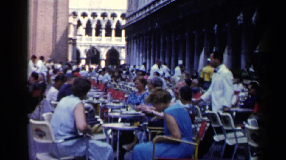 Mid-century Venice outdoor cafe with seated diners, white-jacketed waiters, red chairs, and arched stone architecture in the background.