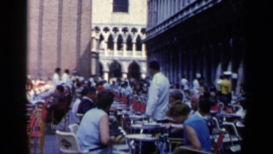 1960 Venice cafe view showing seated patrons, a waiter in a white jacket, rows of red chairs, and arched buildings beyond.