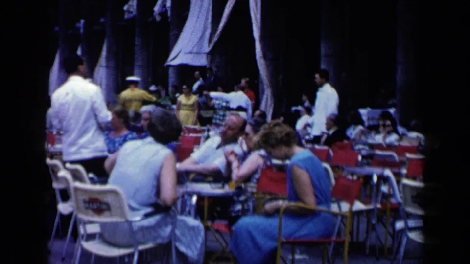 Venice outdoor cafe in 1960 with women in sleeveless blue dresses, nearby waiters, umbrellas, and crowded tables under a shaded colonnade.