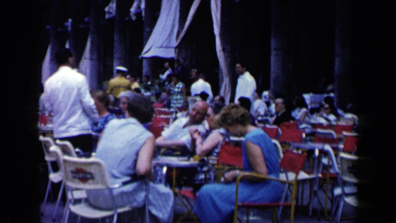 Outdoor café patrons sit at tables under draped canopies with waiters moving through the scene.