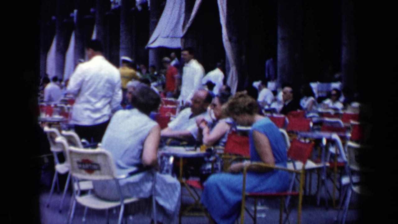 Busy café seating area with patrons at tables, red chairs, white chairs, and servers nearby.