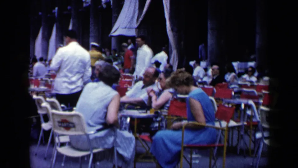 Outdoor café in Venice with patrons seated in period clothing, waiters standing nearby, and rows of red and white chairs.