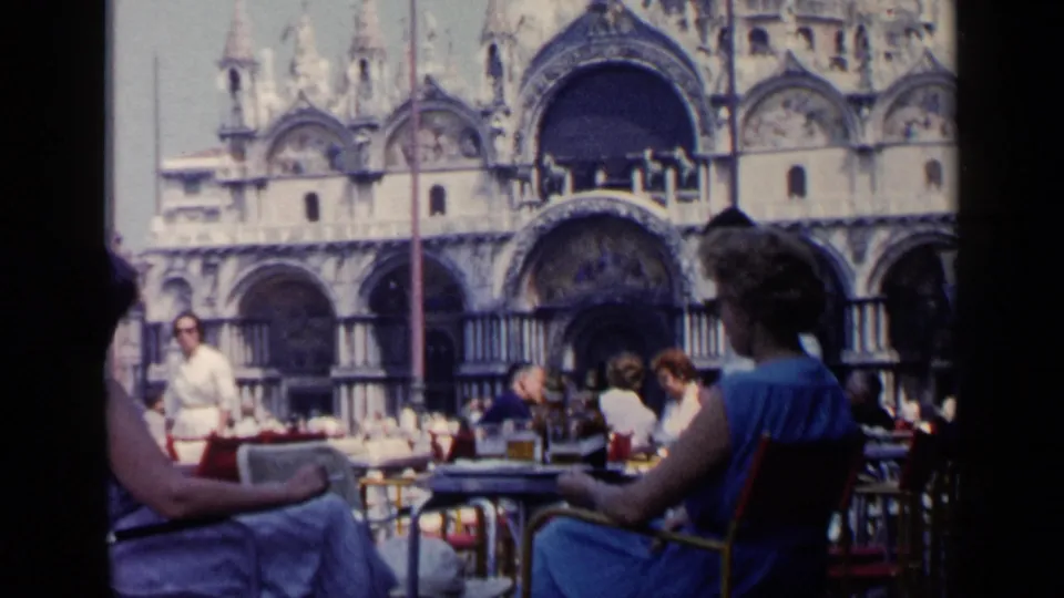 Venice café foreground with seated visitors and a waiter, with the ornate Basilica di San Marco façade in the background.