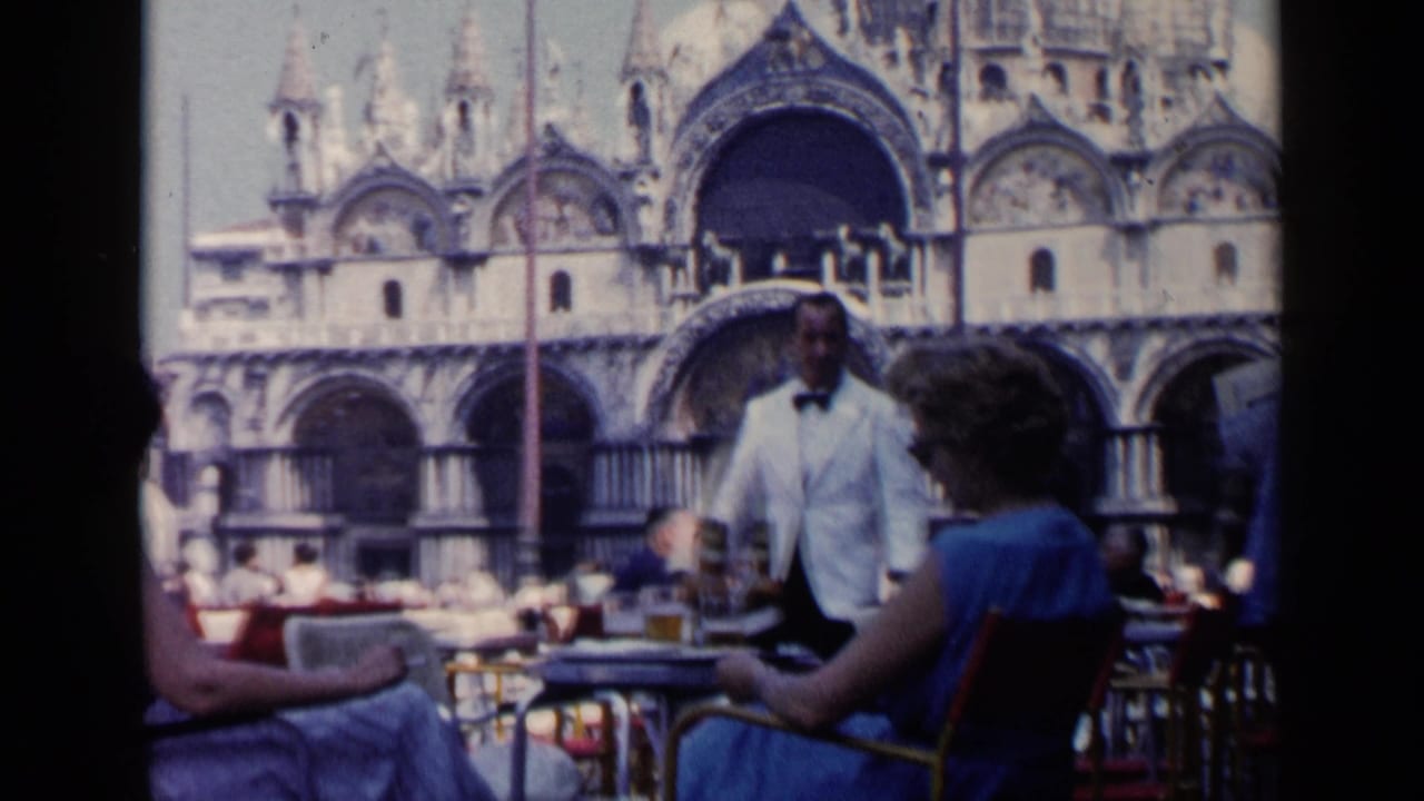 1960 Venice café scene with seated patrons, a white-jacketed waiter, tables, drinks, and St. Mark's Basilica in the background.