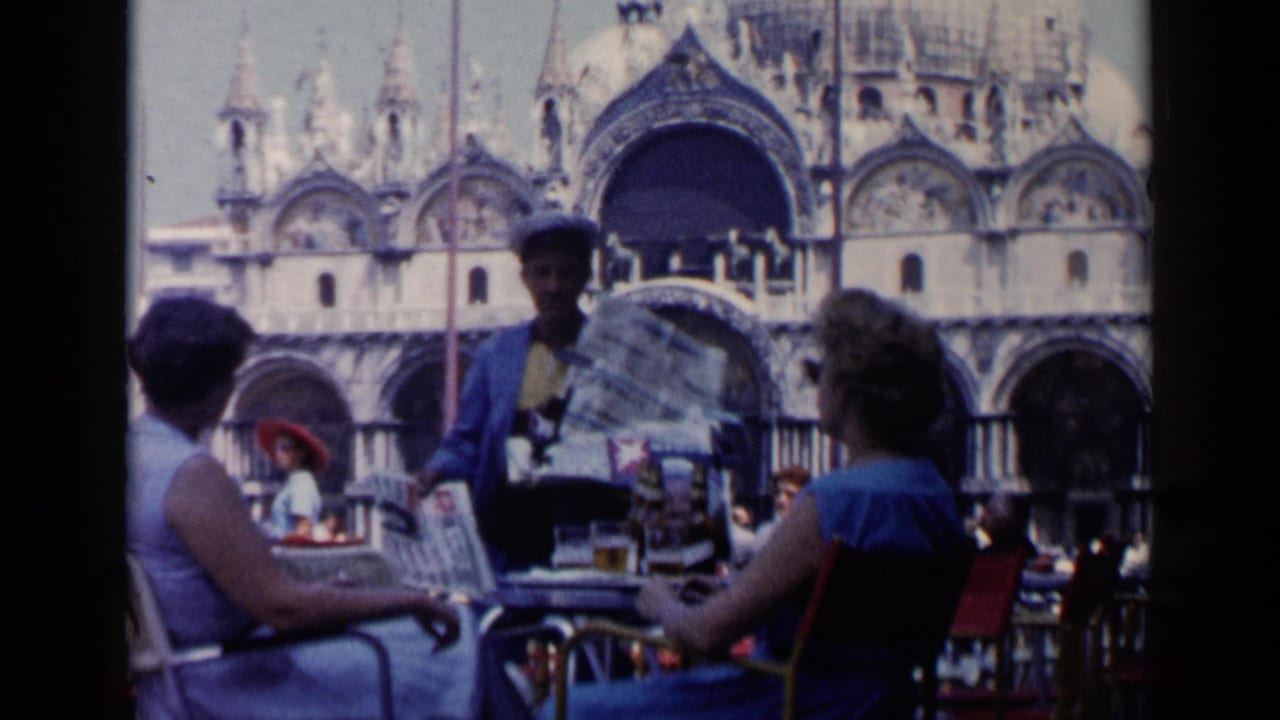 1960 Venice outdoor café view with a newspaper vendor offering papers to seated patrons in front of St. Mark's Basilica.