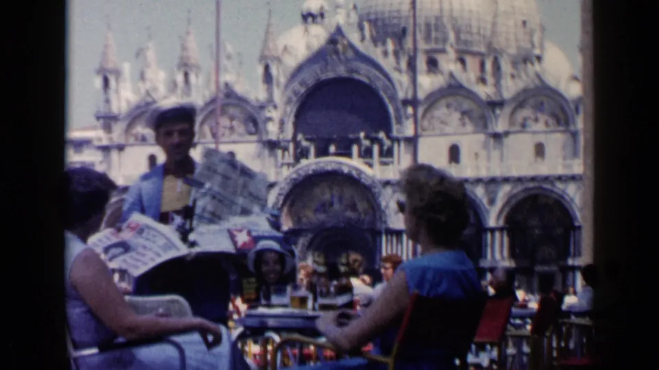 Venice café patrons sit at tables as a blue-jacketed newspaper vendor displays papers, with St. Mark's Basilica behind them.