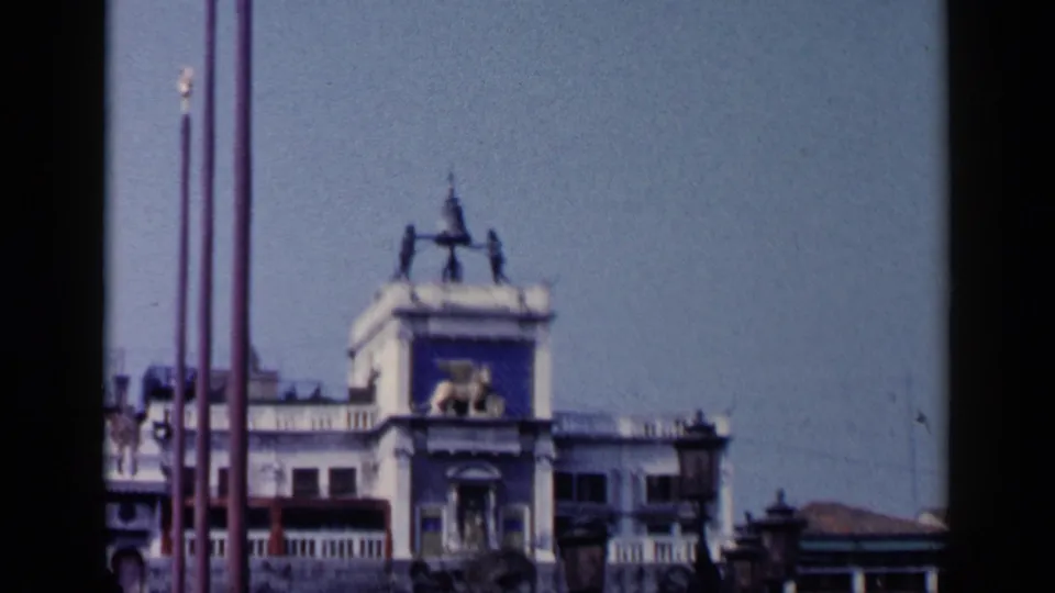 Venice street view with tall flagpoles, a rooftop monument, surrounding buildings, and clear sky in a brief travel cutaway.