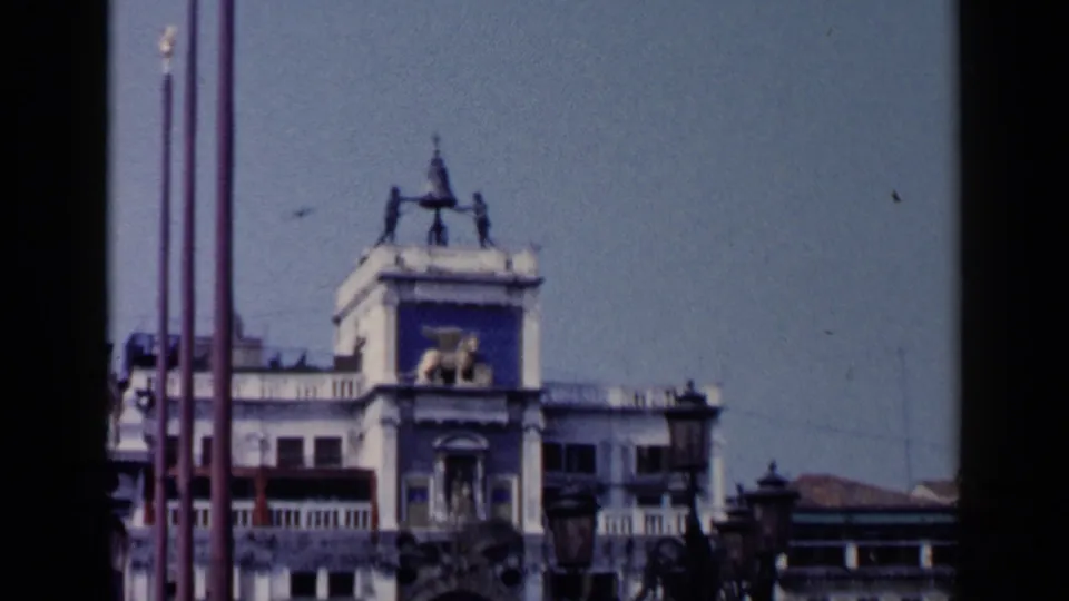 1960 8mm view of the upper St. Mark's Square clock tower in Venice, with a winged lion sculpture and vertical flagpoles.