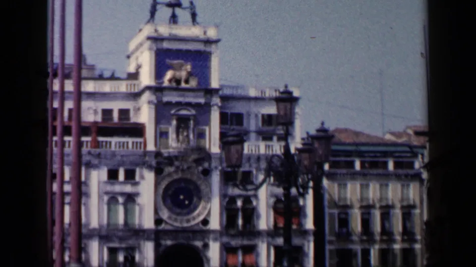 A wider 1960 8mm view in Venice shows the clock tower, large clock face, lower arch, flagpoles, and adjacent facades.