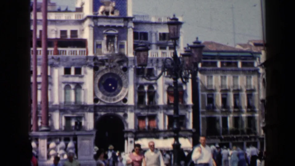 1960 Venice home-movie frame of the St. Mark's Square clock tower above visitors walking through the piazza.