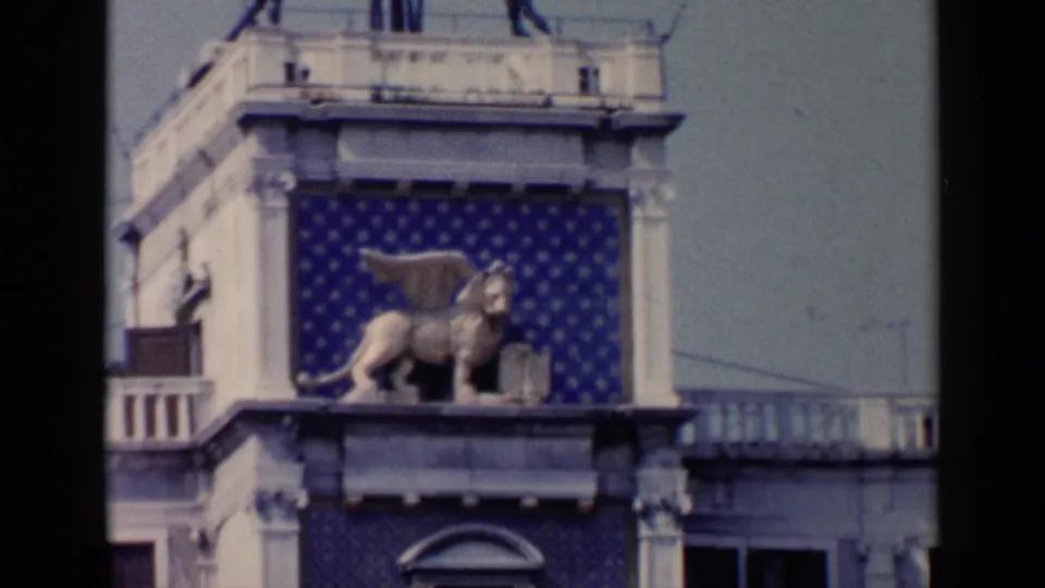1960 Venice frame focused on a winged lion statue set before blue patterned panels and pale architectural trim.