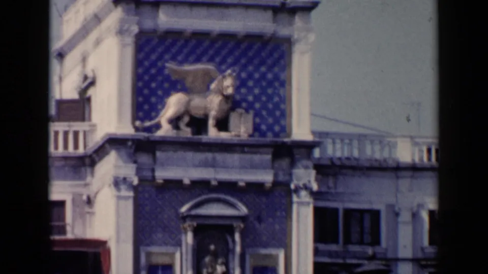 1960 Venice frame showing the winged lion statue above arched window details, blue panels, and adjacent building edges.