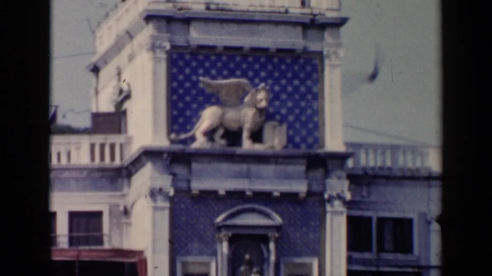 Visitor-shot 1960 Venice frame showing a winged lion-like statue above columns, windows, and a blue patterned wall.