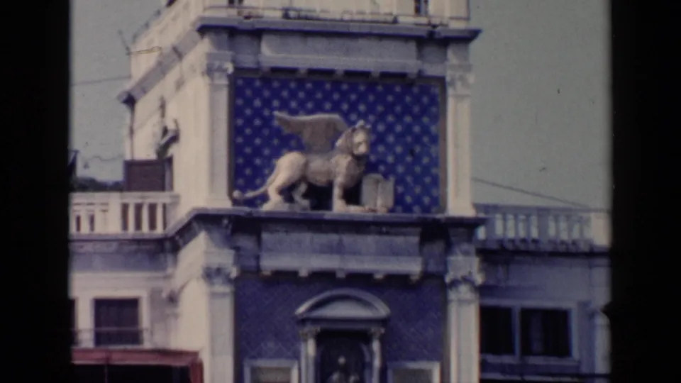 1960 Venice 8mm frame of the winged lion-like sculpture, blue patterned tile, pale stonework, and surrounding windows.