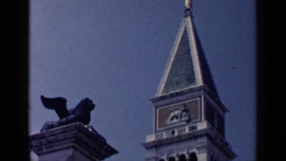 1960 Venice 8mm frame showing a column-top winged lion silhouette beside the bell tower and green-tiled spire against blue sky.