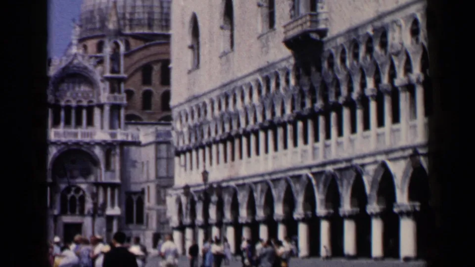 1960 Venice 8mm frame showing St. Mark's Square with basilica architecture, long arcades, and visitors walking below.