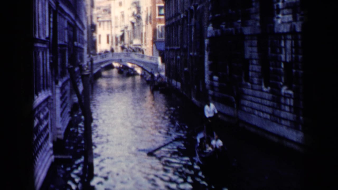 Gondola on a narrow canal approaching a small arched bridge between tall buildings.