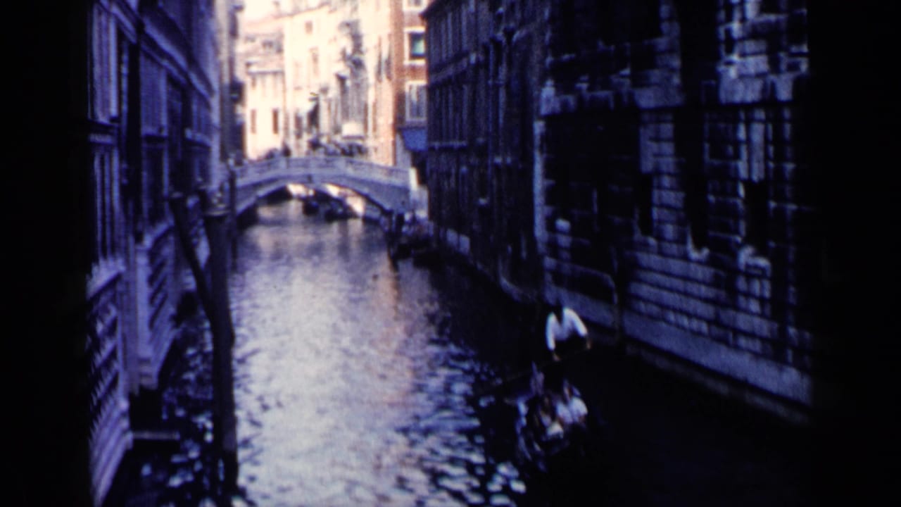 1960 Venice footage frame with a gondolier and tourists passing through a dark canal corridor beneath tall facades.