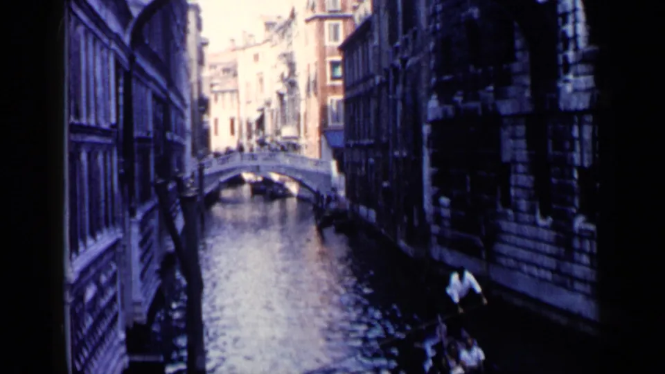 A 1960 Venice home-movie frame looks down a canal as a gondola carries tourists past weathered buildings toward a bridge.