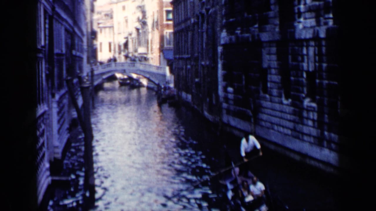 Gondolier rows a gondola carrying passengers along a narrow canal beside stone buildings.