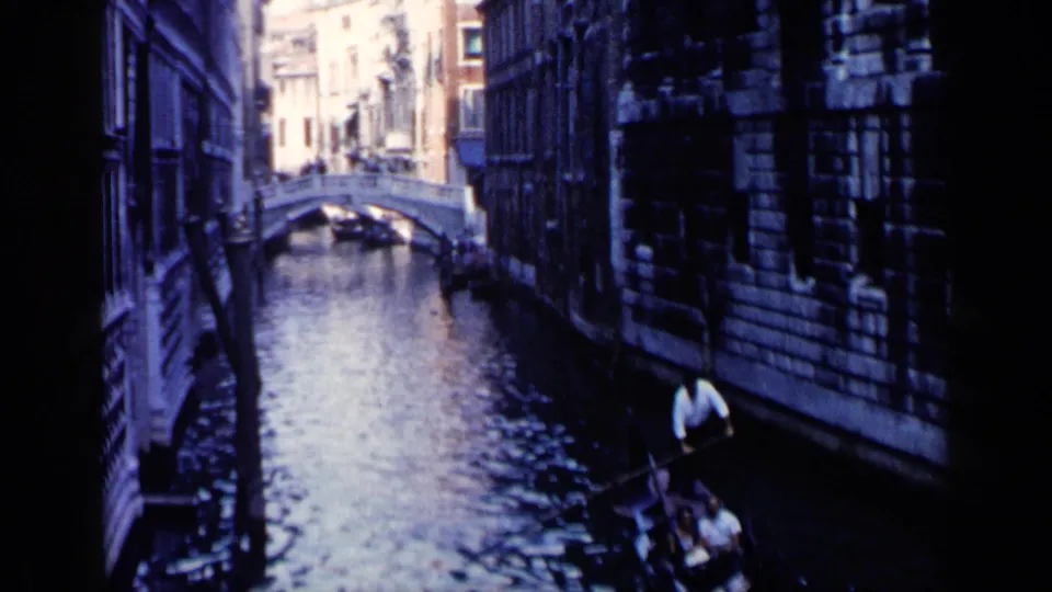 1960 Venice home-movie frame showing a gondolier with passengers in a gondola on a narrow canal between tall stone buildings.