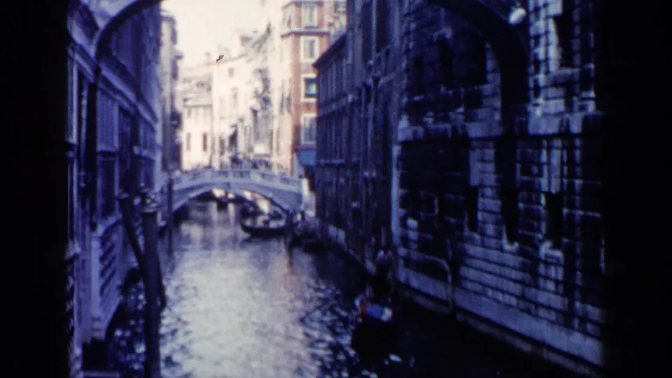 1960 Venice frame with a gondola in a shadowed canal, stone facades on both sides, and a small bridge in the background.