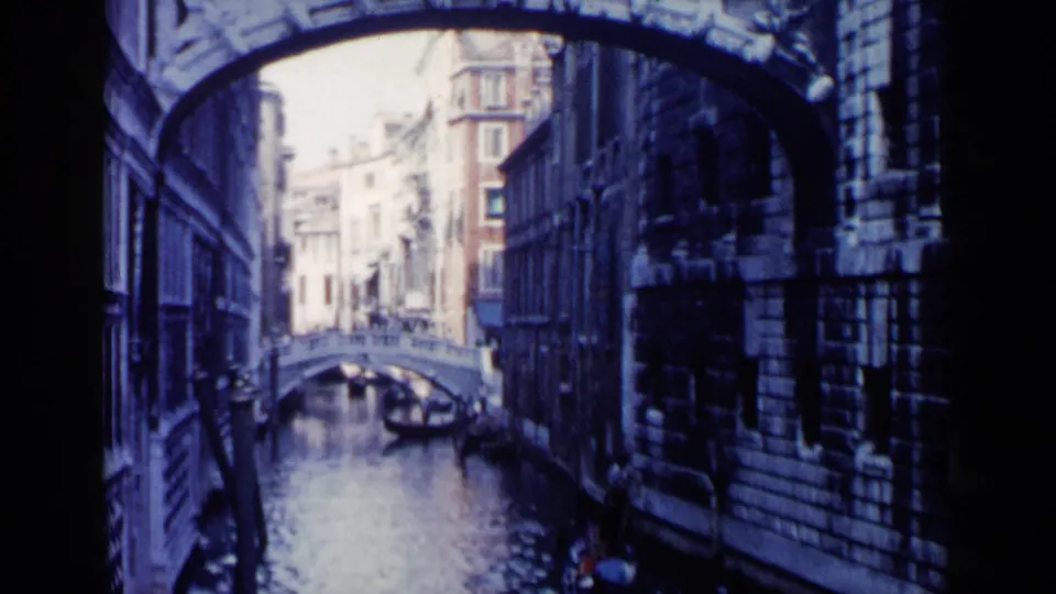 1960 Venice home-movie frame looking through a dark arch over a canal toward a gondola and bridge among historic buildings.