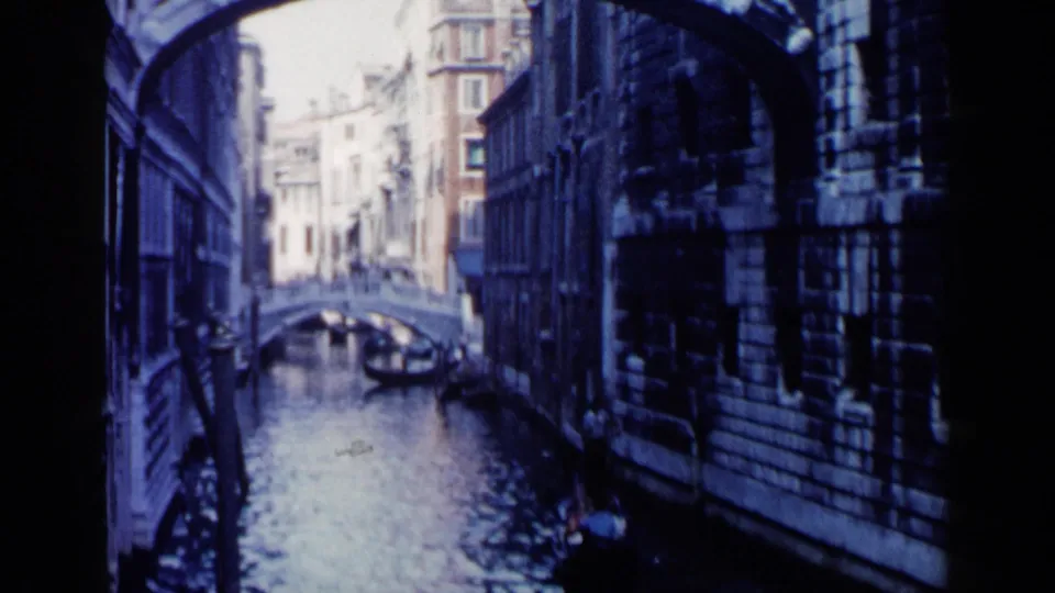 1960 Venice frame showing a gondolier rowing passengers along the right edge of a narrow canal, with a stone bridge ahead.