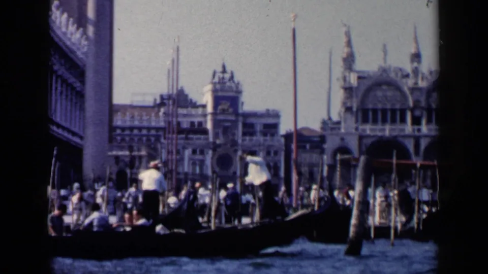 1960 Venice travel frame with a gondolier in a light shirt guiding passengers past arched waterfront architecture and mooring poles.