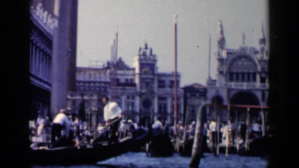 1960 Venice 8mm frame showing multiple gondolas near the waterfront, with visitors, poles, and soft-focus landmark architecture.