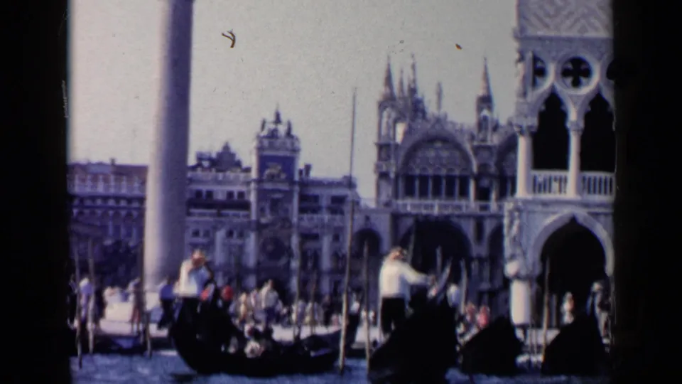 1960 Venice frame with a gondola in the foreground, a standing gondolier, seated passengers, crowds, and columned waterfront architecture.