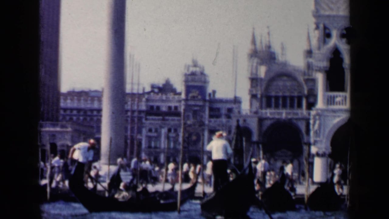 Gondolas and standing gondoliers pass in front of columns and ornate waterfront buildings.