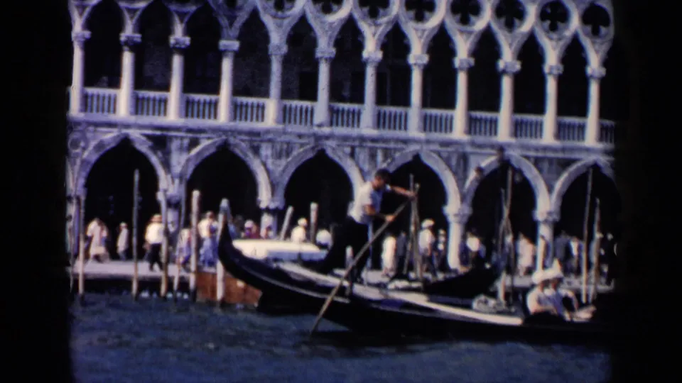 1960 8mm Venice frame showing a gondolier rowing a black gondola beside arched arcades and waterfront pedestrians.