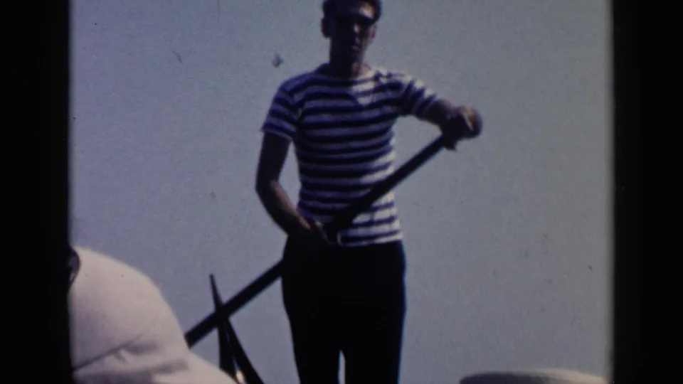 Close 1960 Venice home-movie view of a striped-shirt gondolier holding an oar, with bright sky and part of a passenger visible.