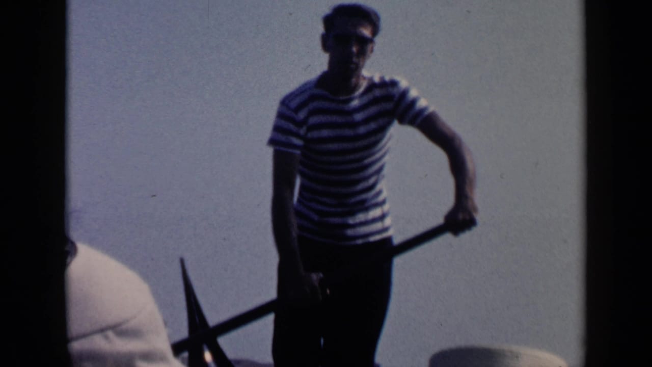 Gondolier in a striped shirt stands with an oar against bright sky.