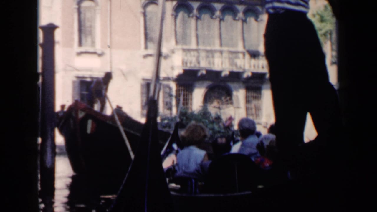 1960 Venice home-movie frame showing gondola passengers, a gondolier, canal water, and ornate building facades beyond dark foreground shapes.