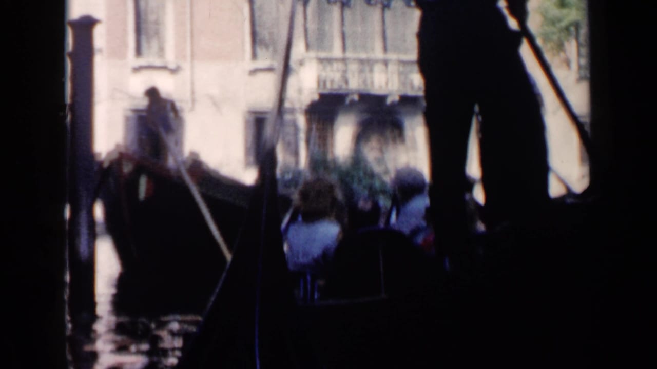 A gondola moves past passengers and pale waterfront buildings, partly obscured by dark foreground shapes.