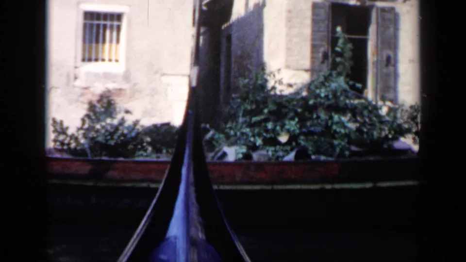1960 Venice home-movie view of a gondola bow in shadow, canal water, plants, and older masonry with barred and shuttered windows.