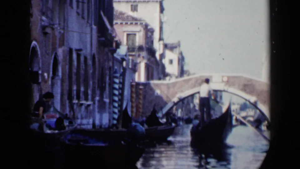 1960 Venice canal scene with gondolas, gondoliers, old waterfront buildings, reflections, and a small arched stone bridge in the distance.