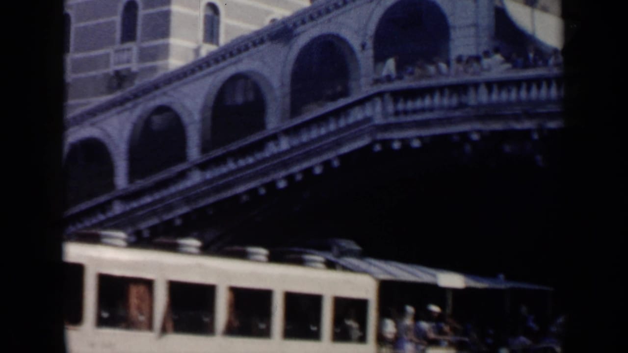 1960 Venice view of the Rialto Bridge with pedestrians above and a canal boat passing below.
