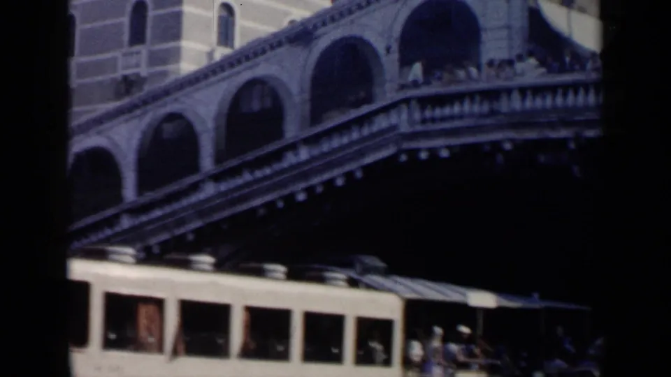 1960 Venice view of the Rialto Bridge with pedestrians above and a canal boat passing below.