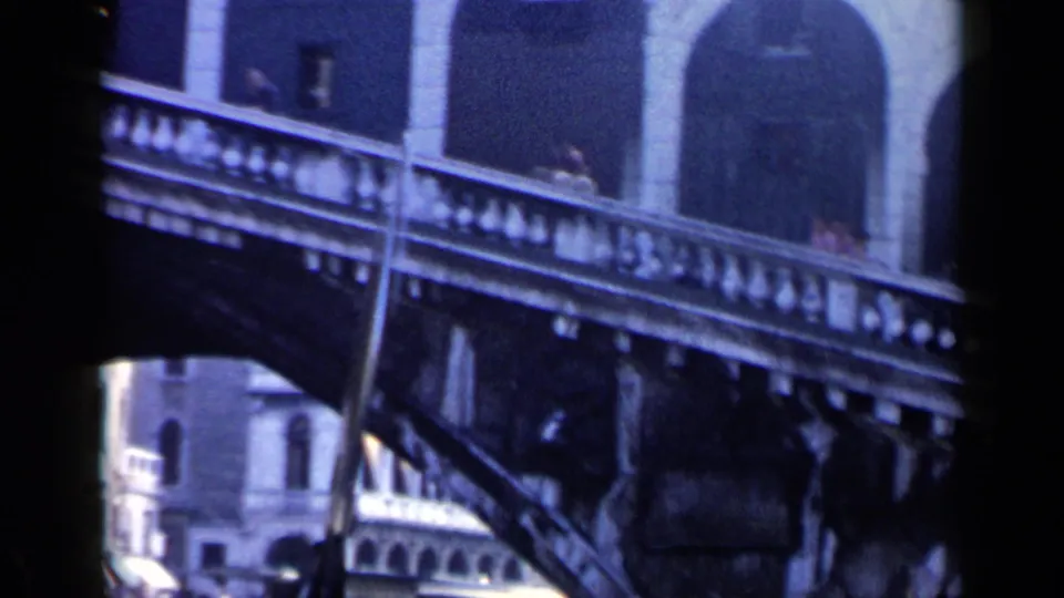 1960 Venice home-movie frame showing the Rialto Bridge structure from a low canal viewpoint, with dark boat edges and soft 8mm color.