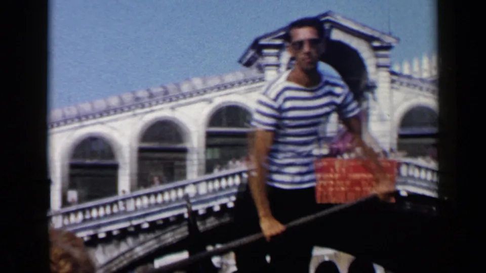1960 Venice frame with a gondolier in striped shirt and sunglasses guiding a gondola near the Rialto Bridge under clear daylight.