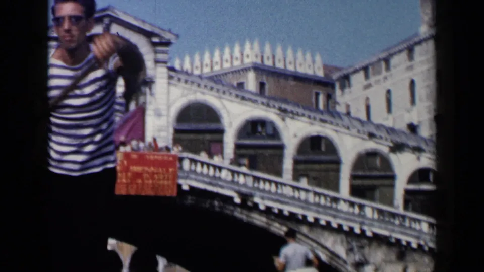 1960 Venice home-movie frame showing a gondolier near red patterned seating, with bridge arches and balustrades in the background.