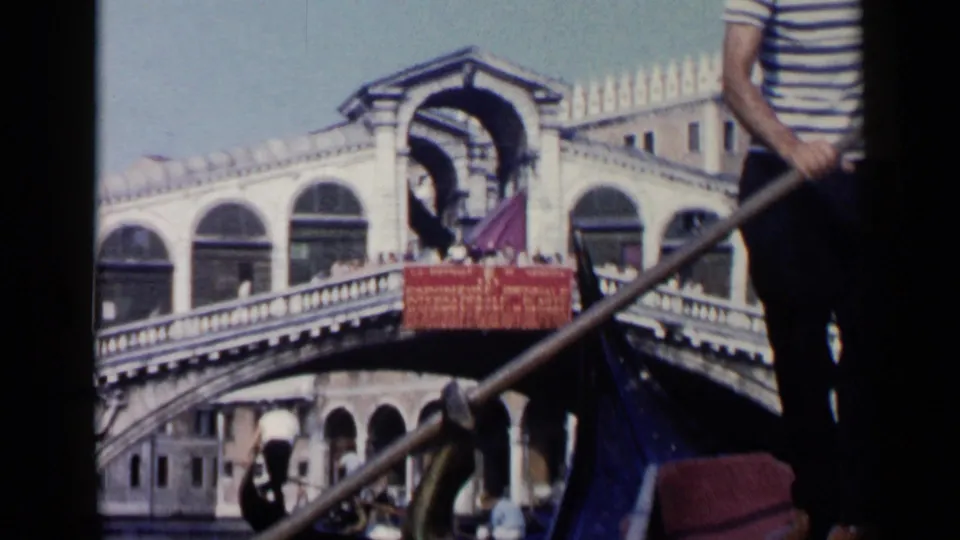 1960 Venice frame showing part of a gondola, red seating, passengers, and the Rialto Bridge receding in the background.