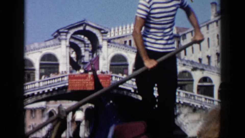 1960 8mm Venice frame showing a striped-shirt gondolier and oar in the foreground with the Rialto Bridge in the background.