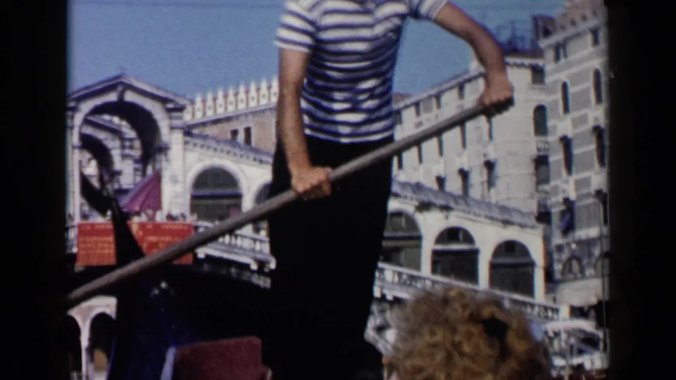 1960 8mm Venice frame with a gondolier rowing near the Rialto Bridge while passengers appear at the lower edge of the gondola view.
