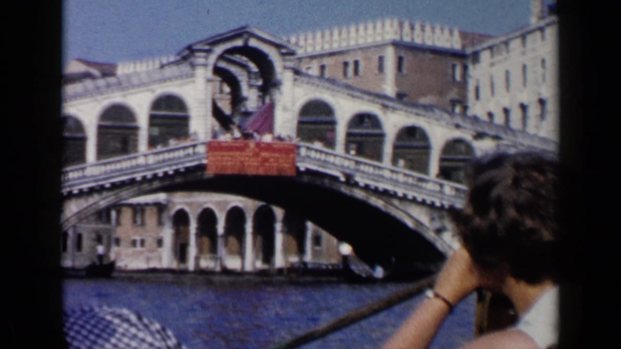 1960 Venice home-movie frame from a gondola, with a passenger in the foreground and the Rialto Bridge across the canal.