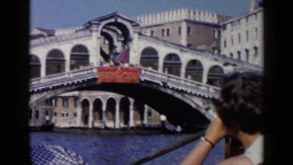 1960 Venice home-movie frame from a gondola, with a passenger in the foreground and the Rialto Bridge across the canal.