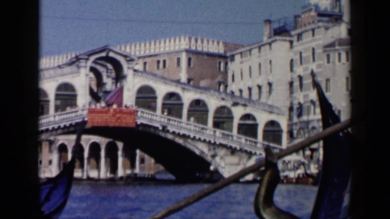 The Rialto Bridge fills the view from a gondola on Venice's Grand Canal, with an oar and gondola bow at the edge.